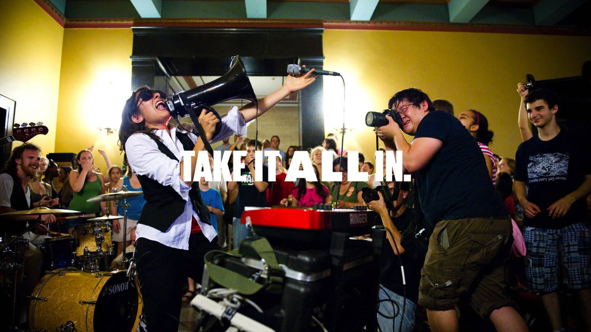 A lively music venue scene with a passionate performer singing into a megaphone. A crowd cheers energetically. Text overlay reads "TAKE IT ALL IN."