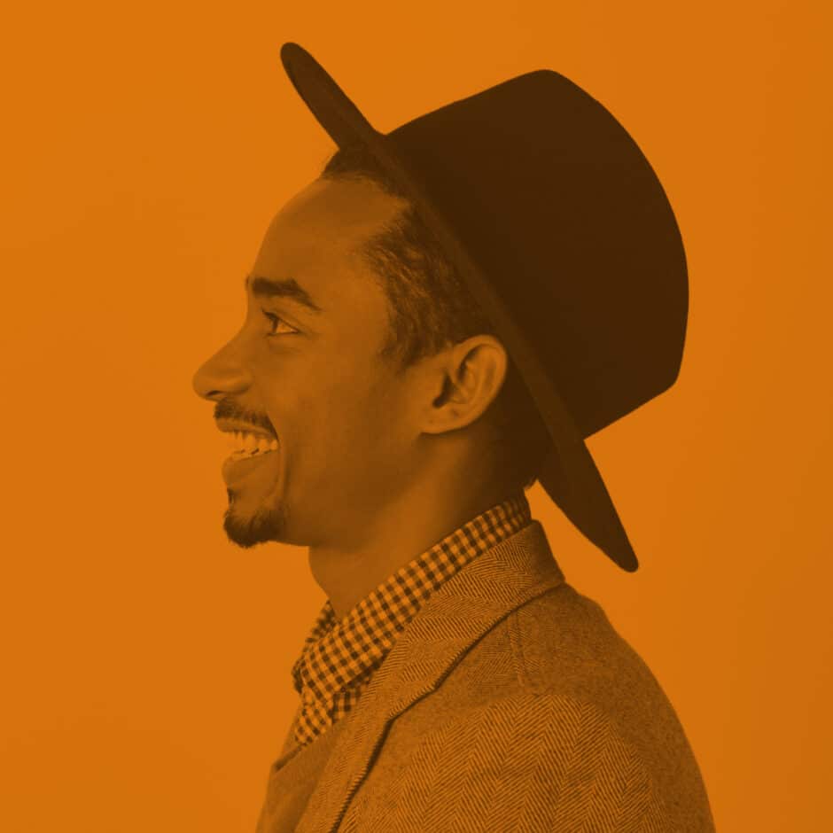 Profile of a smiling man in a hat and blazer against an orange background. The sepia tone adds warmth and a relaxed, vintage feel.