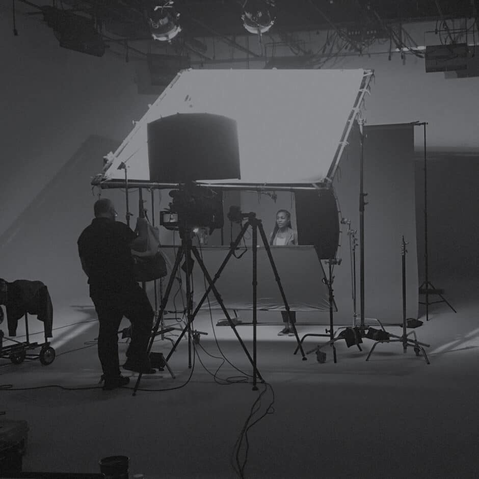 A black and white photo of a professional film set with a large light panel. A person sits behind a desk while a crew member adjusts equipment nearby.