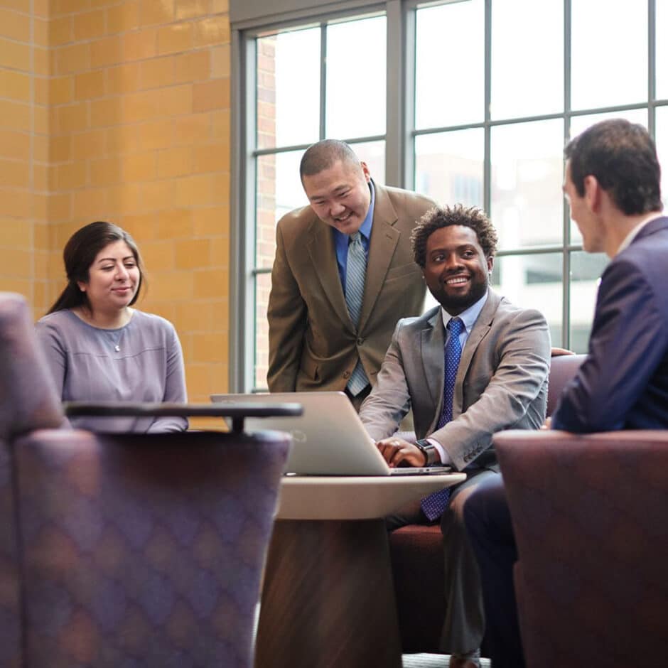 Four professionals in business attire are having a cheerful meeting around a table with a laptop. They sit in a bright room with large windows, exuding collaboration.