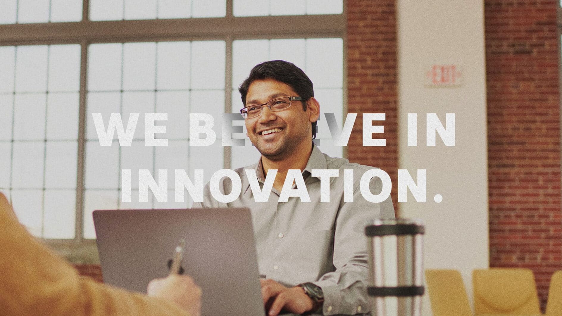 A man smiling while working on a laptop in a modern office setting with exposed brick walls. White text overlays: "We believe in innovation."