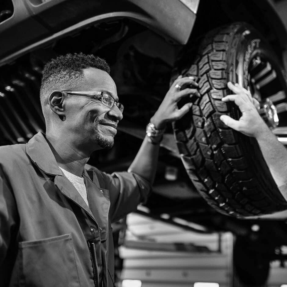 Smiling mechanic in a workshop inspects a car's tire, assisted by another person. The scene conveys teamwork and a positive atmosphere. Black and white.
