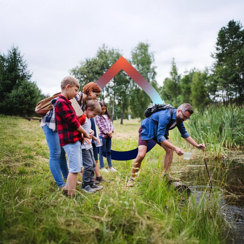 A group of children and a guide gather by a pond, observing him scoop water with a jar. The scene is outdoors, set in lush greenery under a cloudy sky.