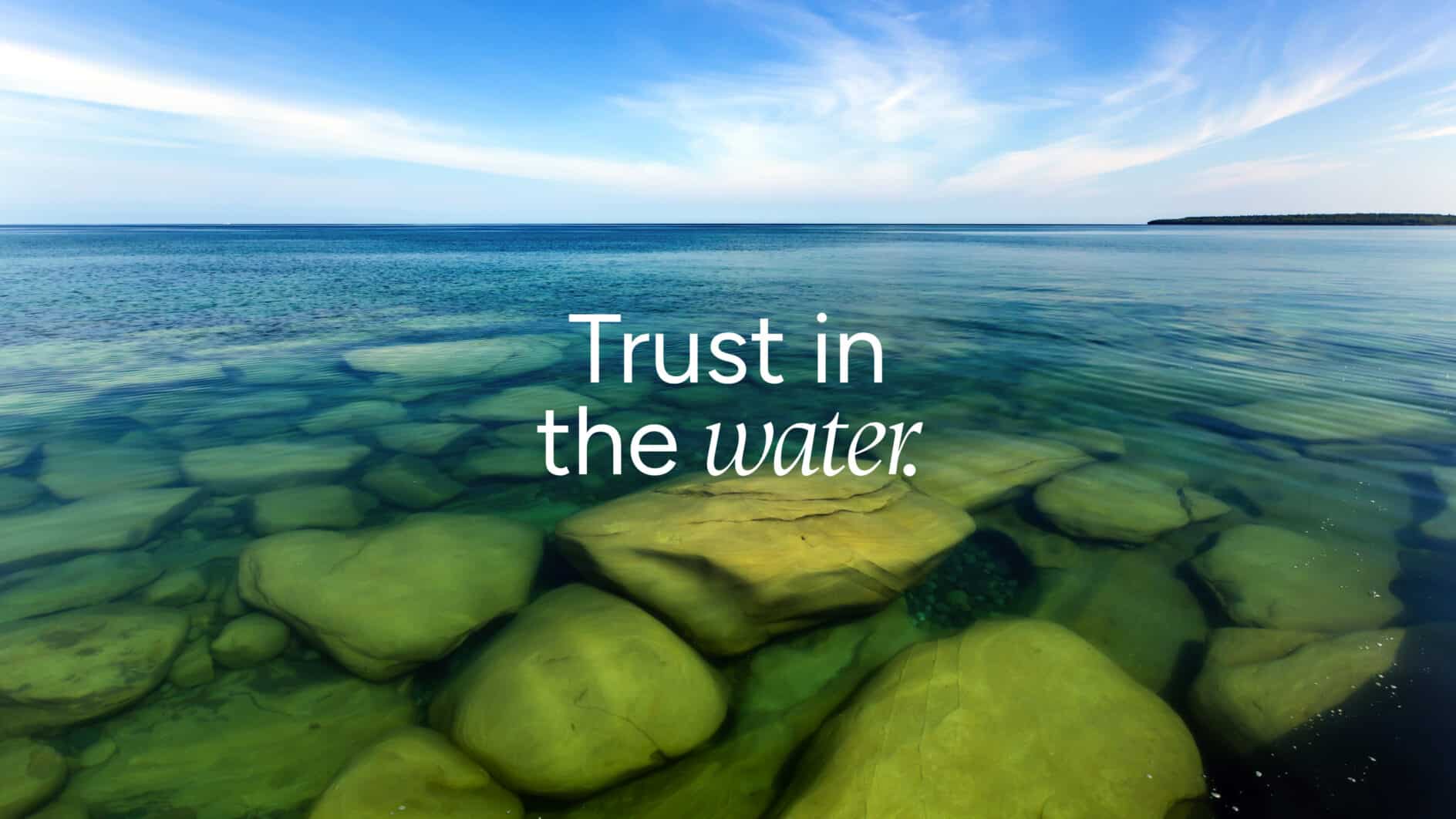 Clear lake water with large submerged rocks, text "Trust in the water" in the center. The horizon shows a serene sky, conveying tranquility and trust.