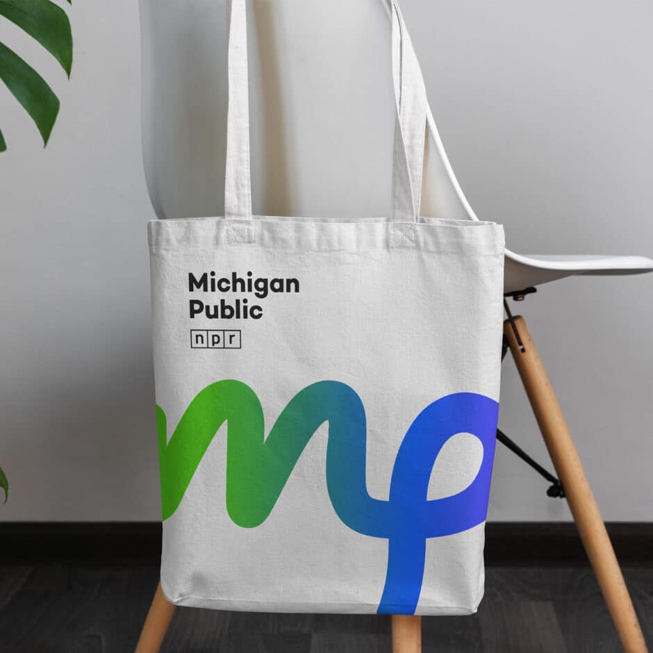 A white tote bag hangs on a chair, featuring the text "Michigan Public NPR" and a large, colorful "mp" design in green and blue. A leafy plant is in the background.