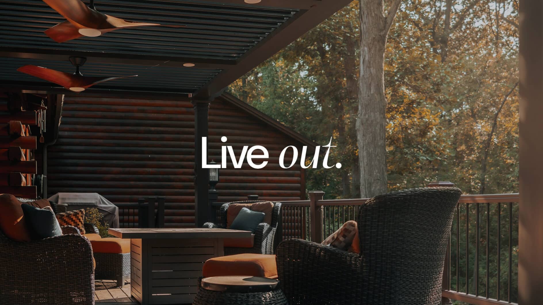 Covered deck with wicker furniture, orange cushions, and a fire table under ceiling fans. Autumn trees surround the cozy, inviting outdoor space. Text reads "Live out."