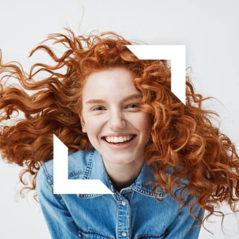 Smiling person with curly red hair wearing a denim shirt, against a white background. The image evokes happiness and vibrancy, enhanced by overlayed white geometric shapes.