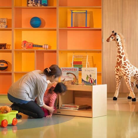 A woman helps a child in a colorful playroom with orange and yellow shelves holding toys and books. A toy giraffe stands nearby, creating a joyful atmosphere.