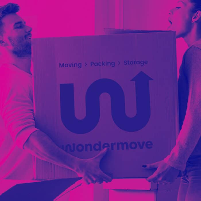 Two people lift a large cardboard box labeled "wondermove" with "Moving > Packing > Storage." The image has a purple-pink gradient, conveying teamwork.