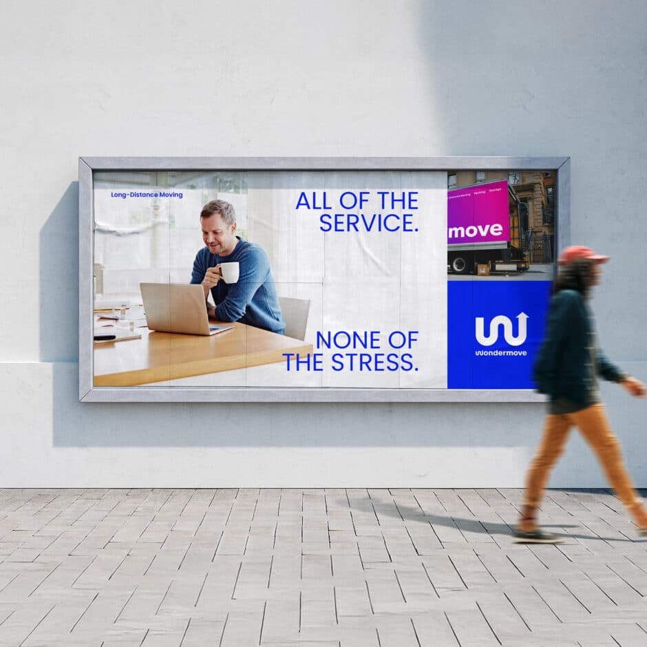 A billboard features a man in a blue sweater working on a laptop at a wooden table, holding a cup. Text reads, "All of the service. None of the stress." A moving truck is displayed with the brand "Wondermove." A person walks by in the foreground.
