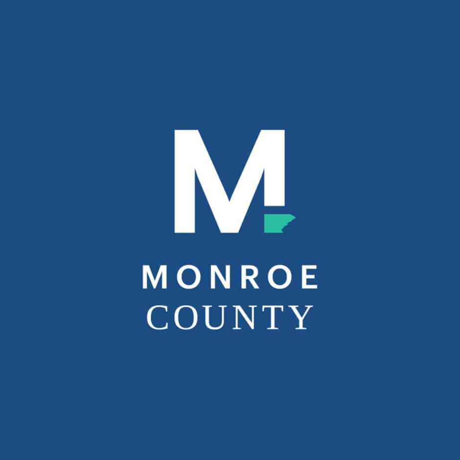 White "M" with teal county outline on blue background, reading "Monroe County" below. Clean, modern design with civic and geographic elements.