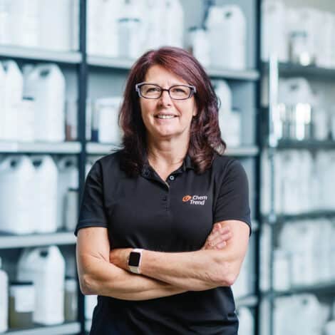 A woman with glasses, wearing a black Chem Trend polo, smiles confidently with arms crossed. Shelves of chemical containers fill the background.