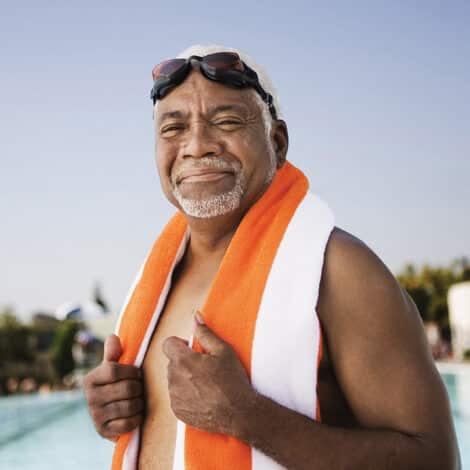 Elderly man with goggles on head, smiling by the poolside. He is wrapped in an orange and white towel, exuding a relaxed and content mood under a clear sky.