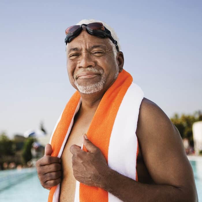 Elderly man with goggles on head, smiling by the poolside. He is wrapped in an orange and white towel, exuding a relaxed and content mood under a clear sky.