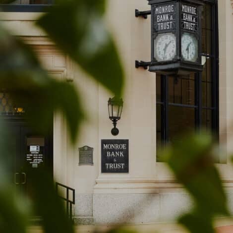 Historic Monroe Bank & Trust building facade with classic signage, a vintage clock, and a black lantern. Green leaves partially obscure the view.