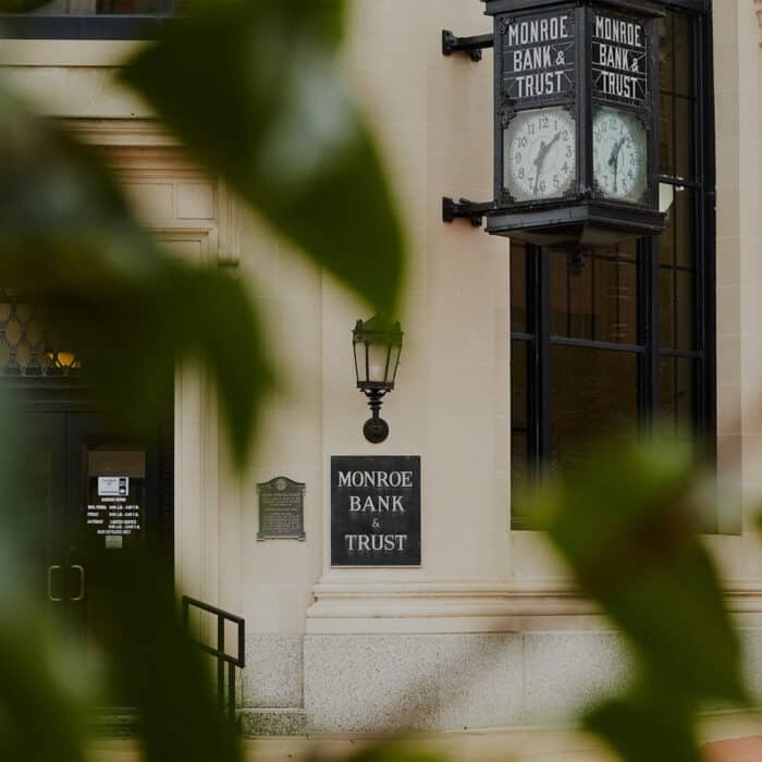 Historic Monroe Bank & Trust building facade with classic signage, a vintage clock, and a black lantern. Green leaves partially obscure the view.