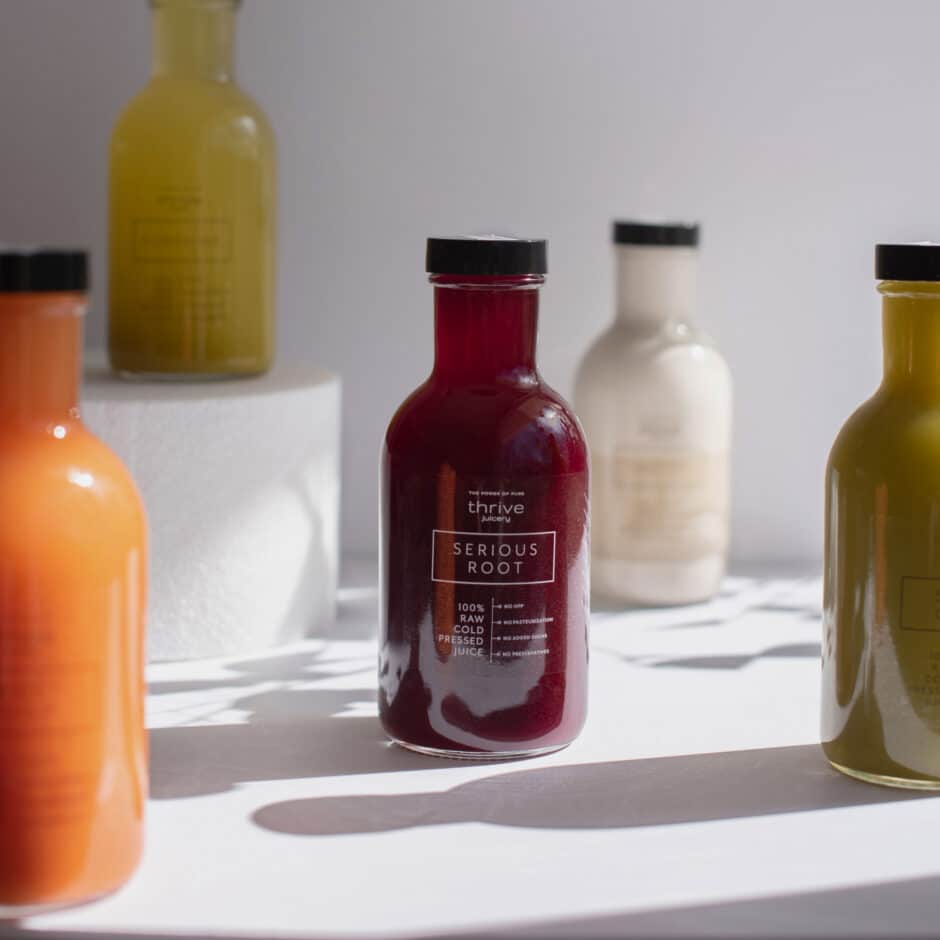 Assorted juice bottles on a white surface with soft lighting. Central red bottle labeled "Serious Root" surrounded by orange, yellow, and cream-colored bottles.