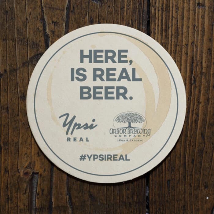A round beer coaster on a wooden surface reads "Here, is real beer." It features logos for Ypsi Real and Arbor Brewing Company. Hashtag #YPSIREAL.