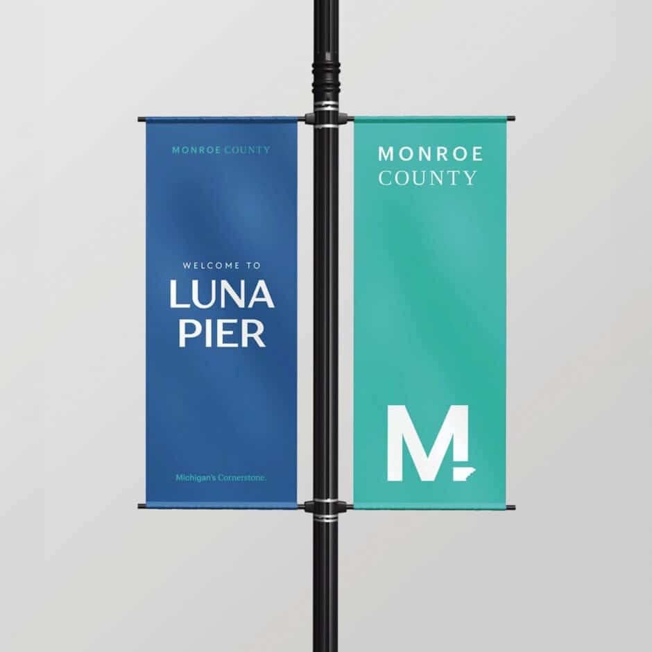 Two vertical banners on a pole; left banner in blue reads "Welcome to Luna Pier, Michigan's Cornerstone," right in teal displays "Monroe County" with a large "M".