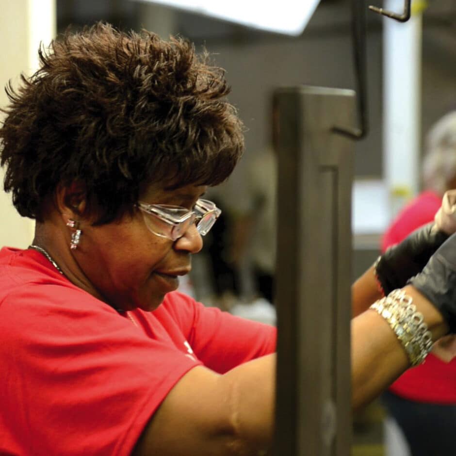 A woman with curly hair, wearing glasses and a red shirt, is focused on working with machinery. Her expression is attentive and concentrated.