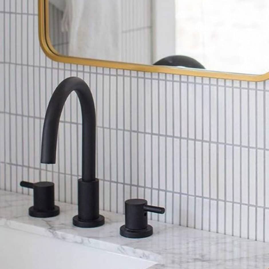 Sleek black faucet with dual handles on a white marble countertop. Set against vertical white tiles and a gold-framed mirror, creating a modern, elegant feel.