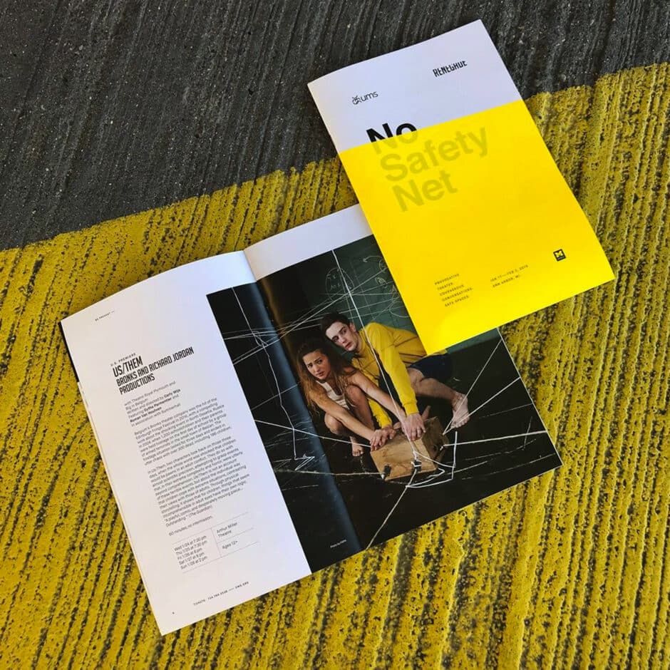 An open magazine with an article about "Us/Them" on a yellow and gray textured surface. A vibrant yellow and white cover titled "No Safety Net" is nearby.