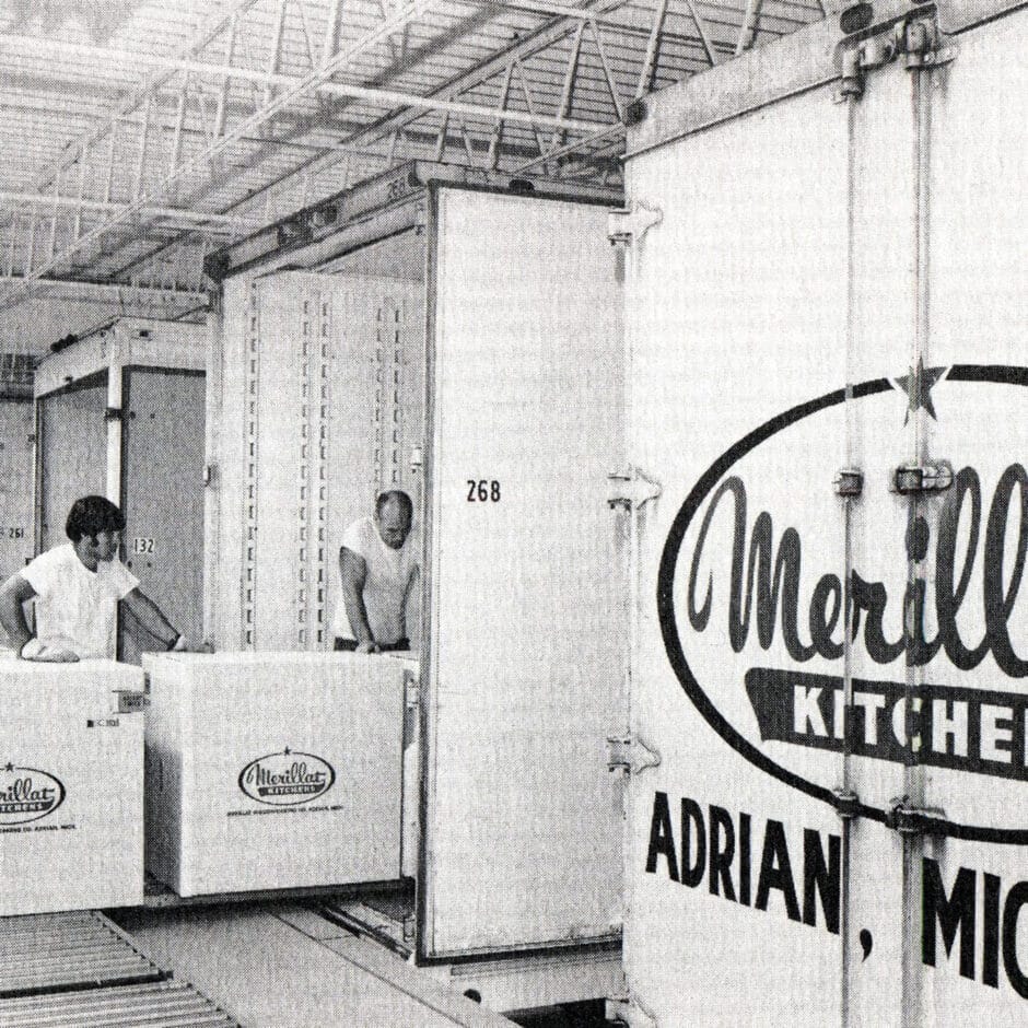 Black and white photo of two men loading large boxes labeled "Merillat Kitchens" into a truck. Industrial setting, focused and busy atmosphere.