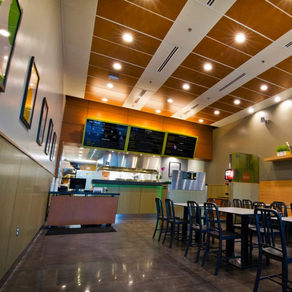 Modern restaurant interior with a high ceiling, bright lighting, and polished floors. Menu boards are displayed above a counter, with tables and chairs in the foreground.
