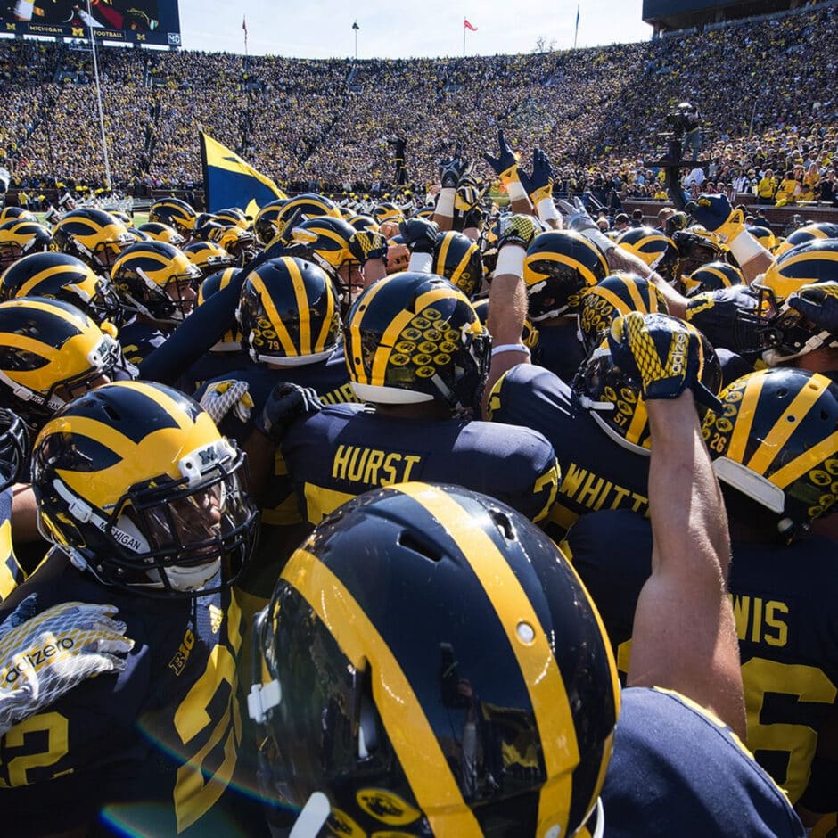 University of Michigan football players wearing navy-and-yellow uniforms and helmets huddle with arms raised in excitement on a sunny day. A large crowd fills the background.