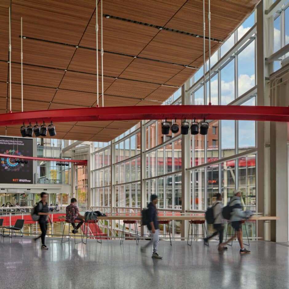 A modern, airy university atrium with large windows, wood ceiling, and red beams. Students walk and sit, creating a lively, dynamic environment.
