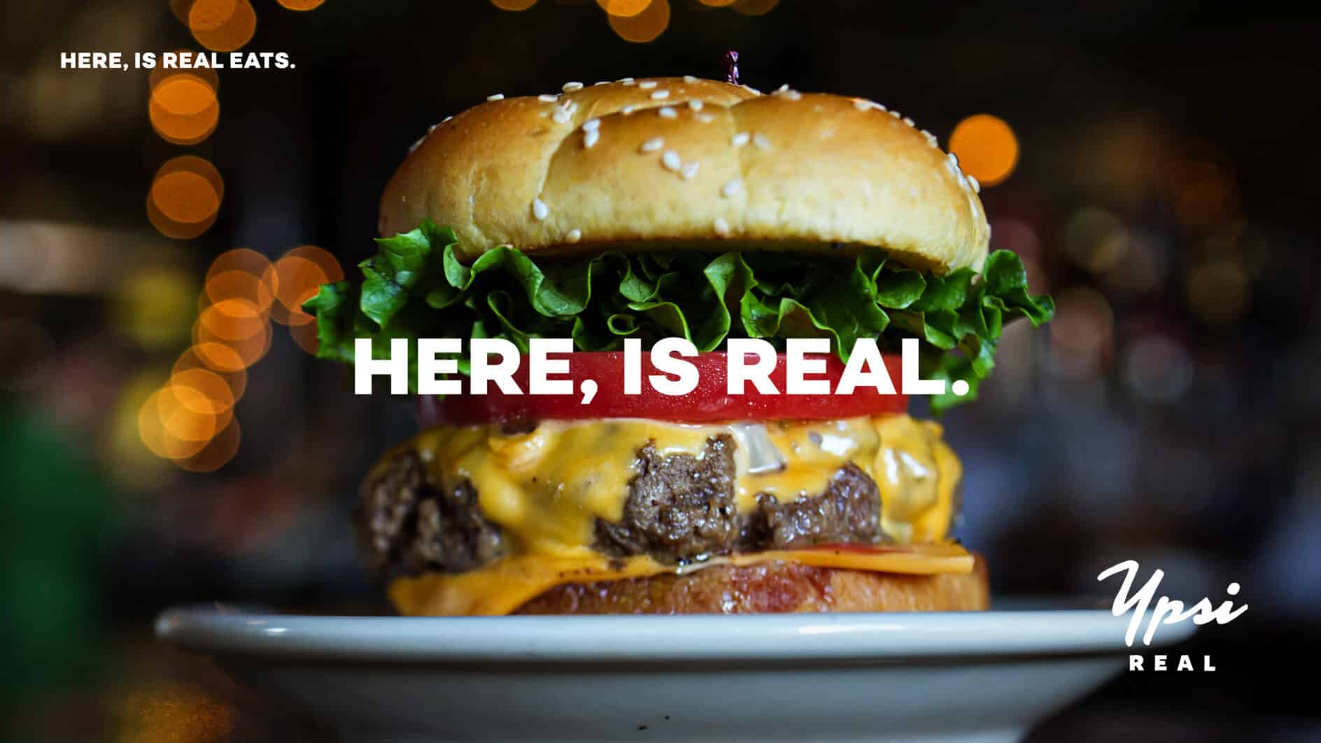 Juicy cheeseburger with lettuce and tomato on a sesame bun, centered on a plate. Background has warm bokeh lights. Text says, "Here, Is Real."