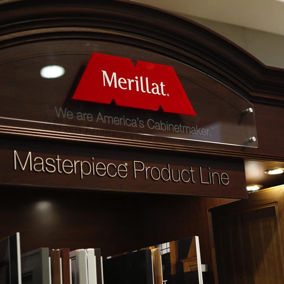 Signage for Merillat's Masterpiece Product Line displayed on a dark wood cabinet. The text reads "We are America’s Cabinetmaker," evoking quality and elegance.