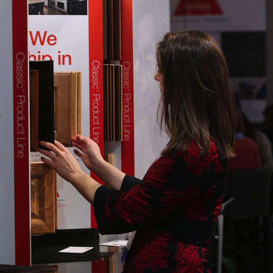 A woman examines wooden cabinet samples in a showroom. She has long hair and wears a black and red outfit. The display is labeled "Classic Product Line."