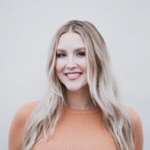 A woman with long blonde hair smiles warmly, wearing an orange sweater against a plain white background. The tone is friendly and inviting.