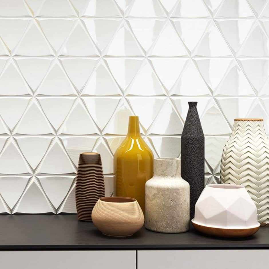 A group of decorative vases and pots in various shapes and textures sits on a black surface. The background features a glossy, geometric white tile wall.