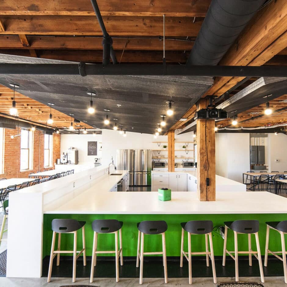 Spacious modern kitchen with wooden beams, bright lighting, and a long white counter with green accents. Black stools and industrial decor add a sleek touch.