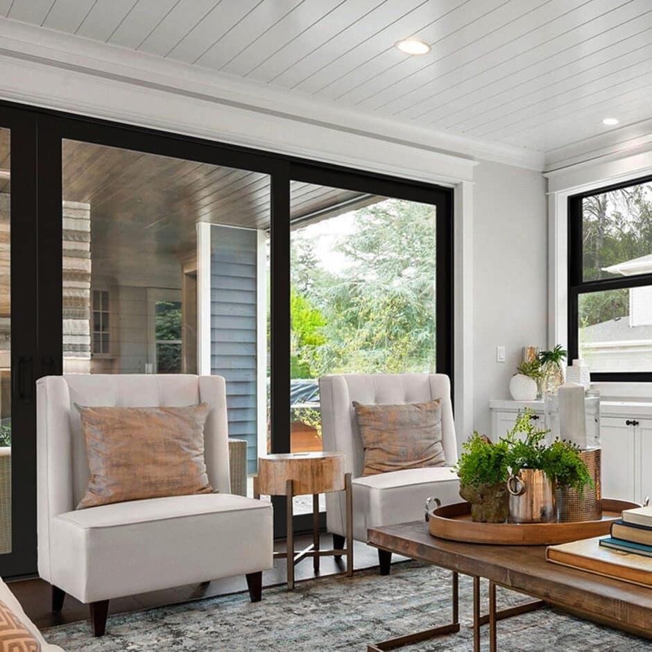 A modern living room features two white armchairs with beige pillows, a wooden side table, and a coffee table with plants, books, and decor by large windows.