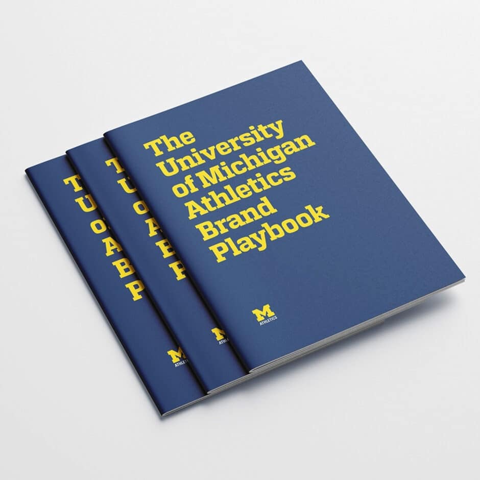 Three blue booklets titled "The University of Michigan Athletics Brand Playbook" with yellow text are stacked. The cover displays the Michigan logo.