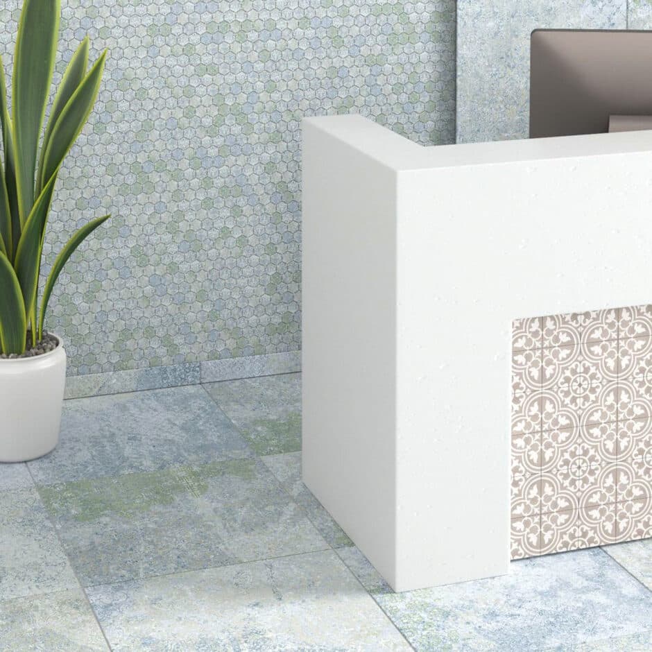 Modern interior with a white reception desk featuring patterned detailing. A potted plant sits beside, with a mosaic wall and tiled floor.