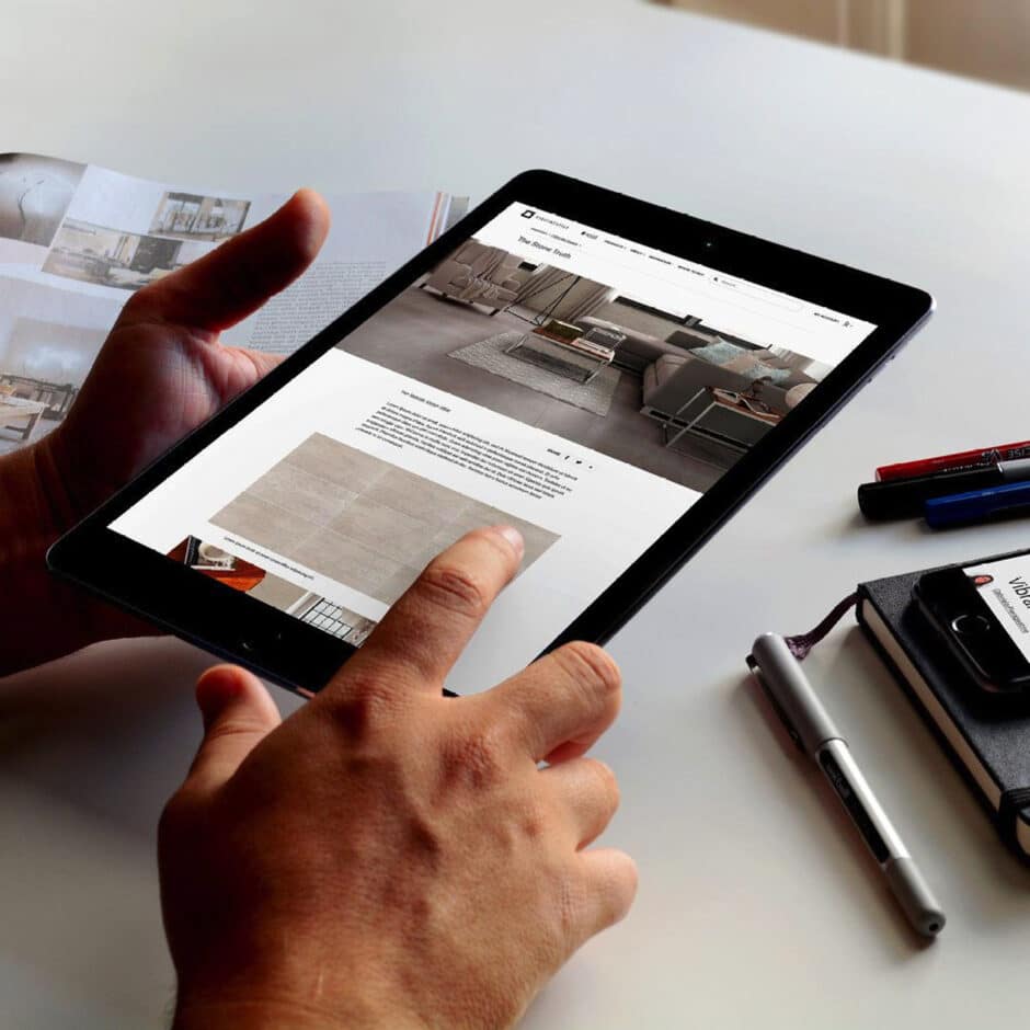 Hands holding a tablet showing a modern interior design website. A person interacts with the screen, while pens and a notebook lie nearby on the table.