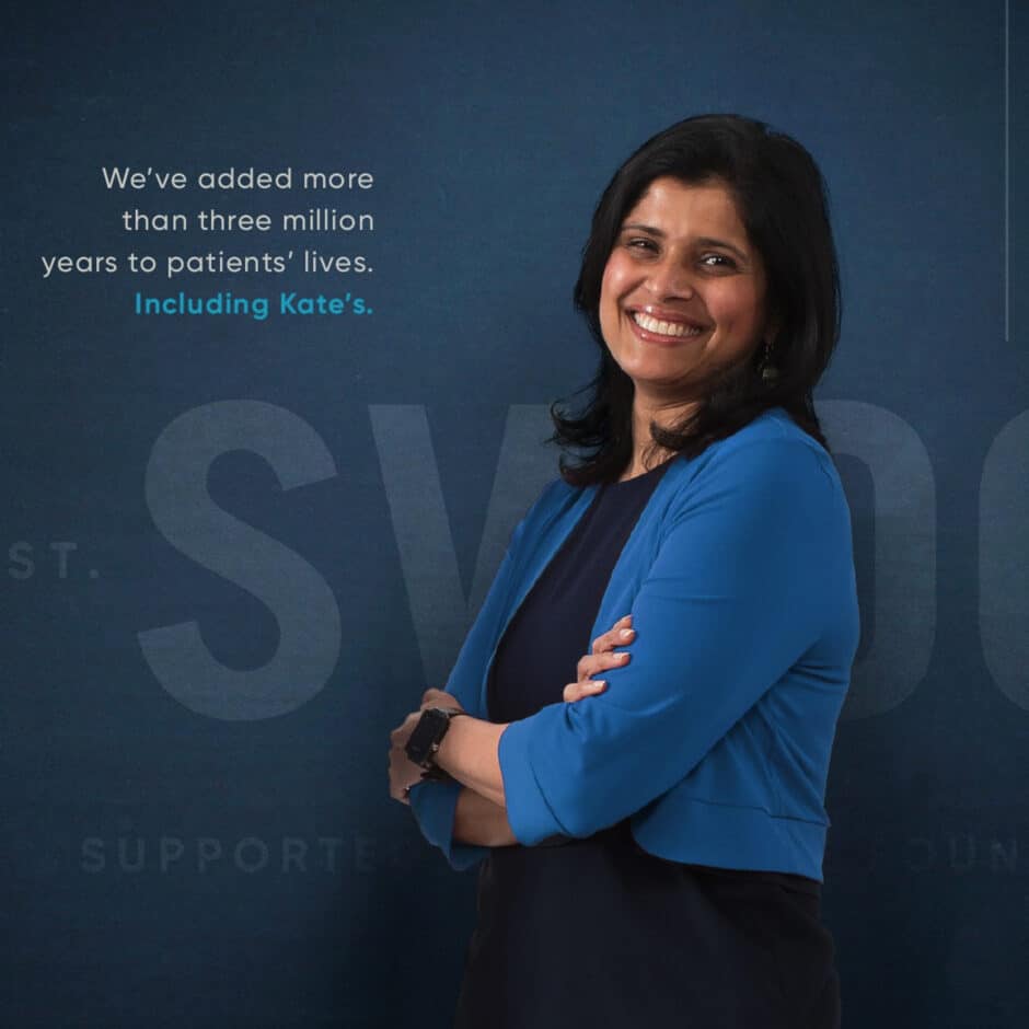 Smiling woman in a blue jacket stands confidently with arms crossed against a dark blue backdrop with text about improving patient lives.