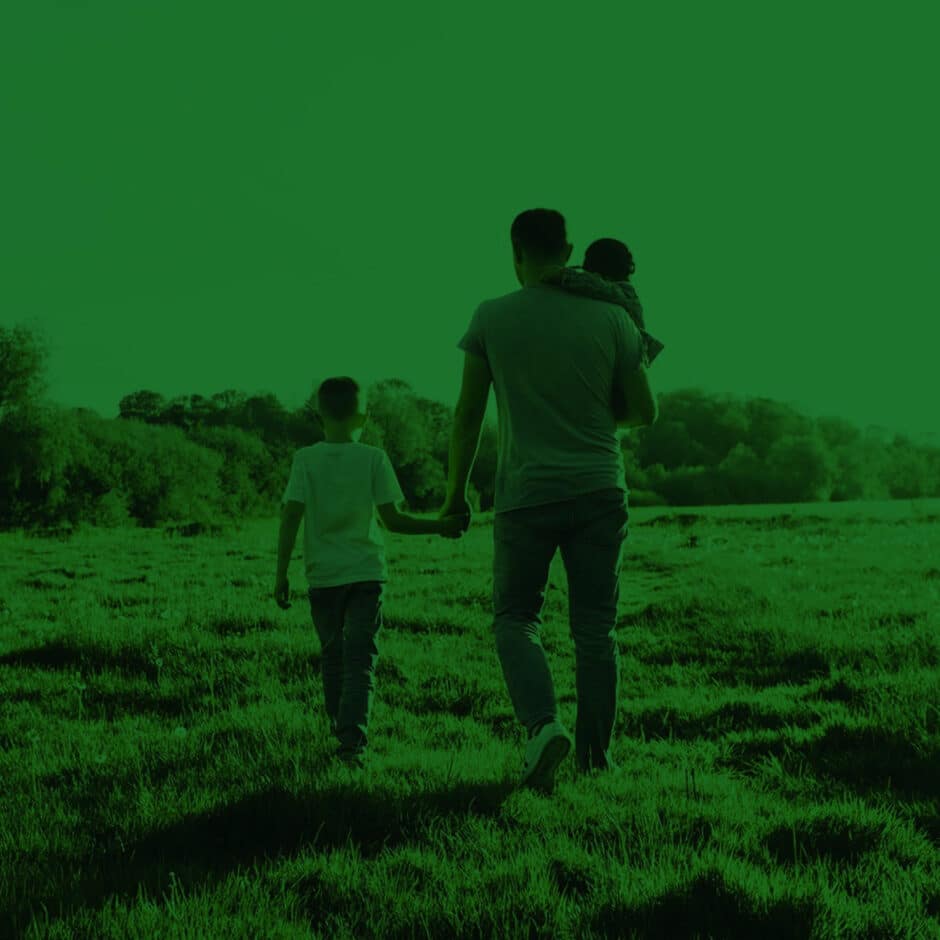 A man walks in a meadow holding hands with a young child while carrying another child in his arm. The scene is tinted green, conveying tranquility.