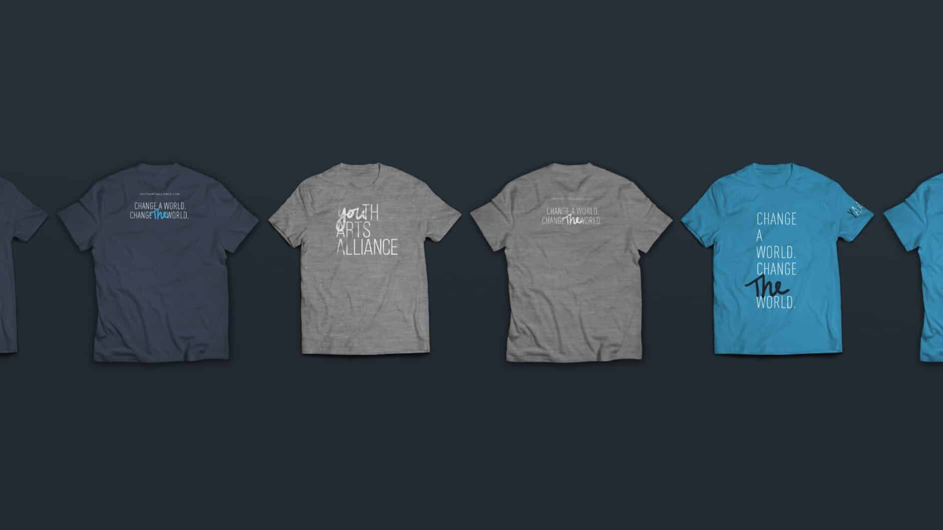 Five T-shirts are displayed horizontally against a dark background. The shirts, in blue and gray, feature motivational texts in various fonts, promoting change and arts.