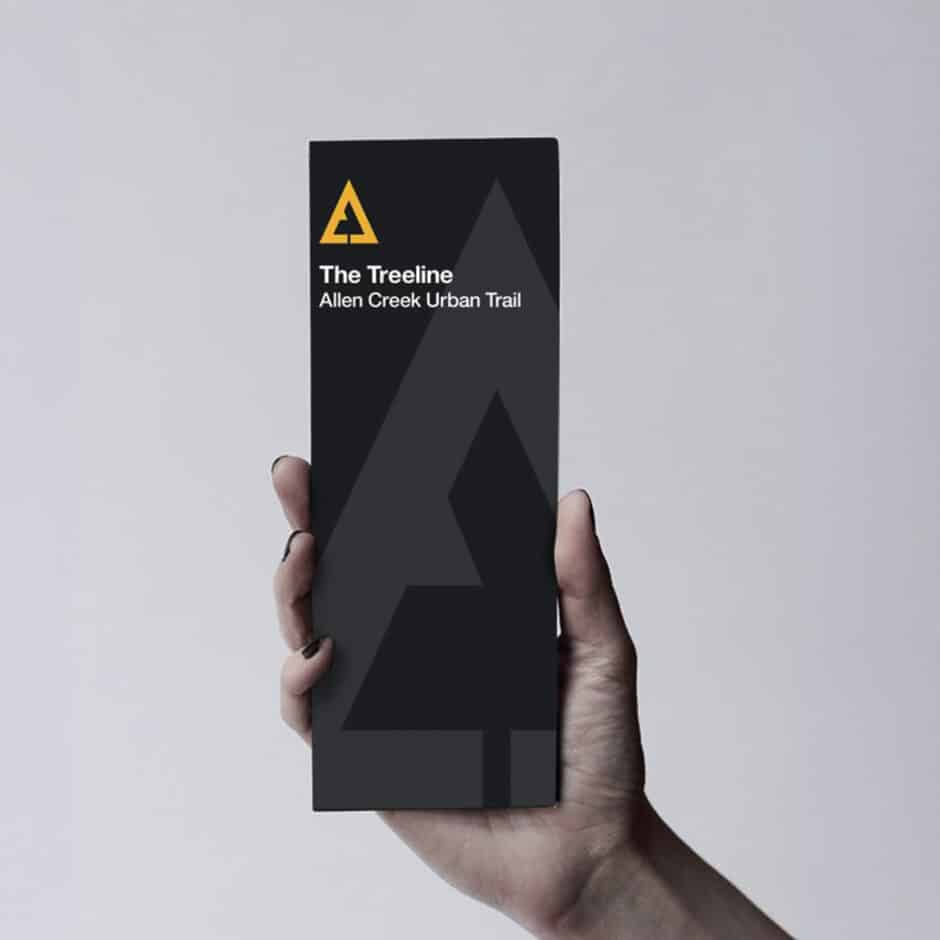 A hand holds a dark gray brochure with a minimalist tree design. The text reads "The Treeline, Allen Creek Urban Trail" under a yellow triangular logo. Clean, modern look.