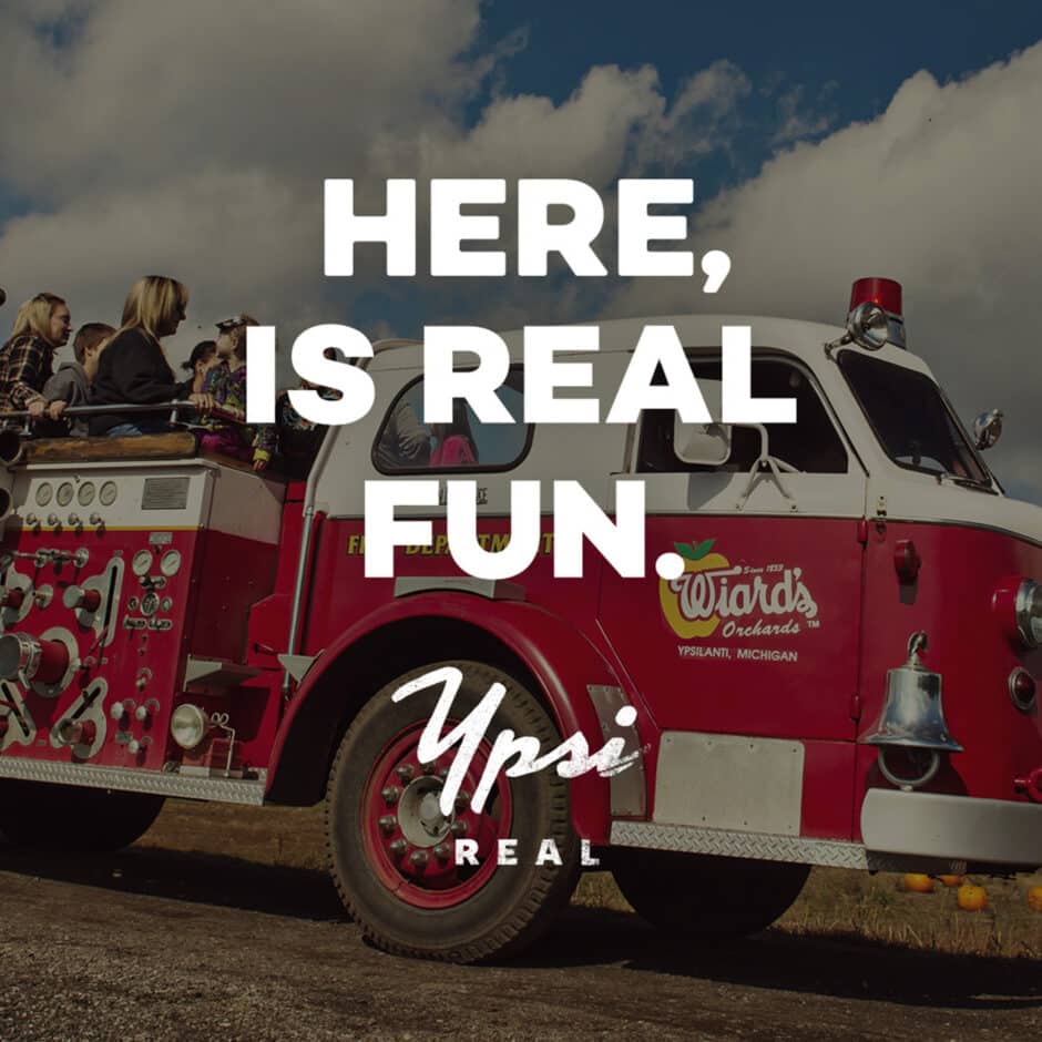 Red vintage fire truck with happy passengers on a sunny day, parked on a dirt path. Bold white text reads: "Here, is real fun. Ypsi Real."