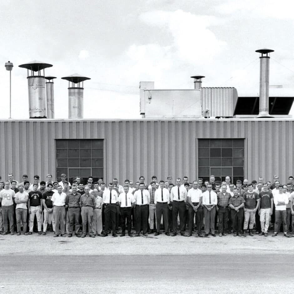 A large group of men stands in front of an industrial building. The men, some in ties and shirts, others in casual attire, convey a formal yet relaxed tone.