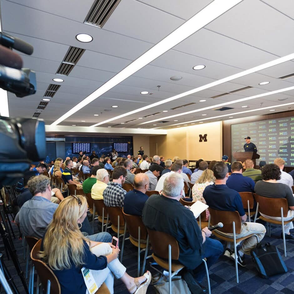 A packed press conference room with a speaker at a podium, branded backdrop, and audience seated in rows. The atmosphere is focused and professional.