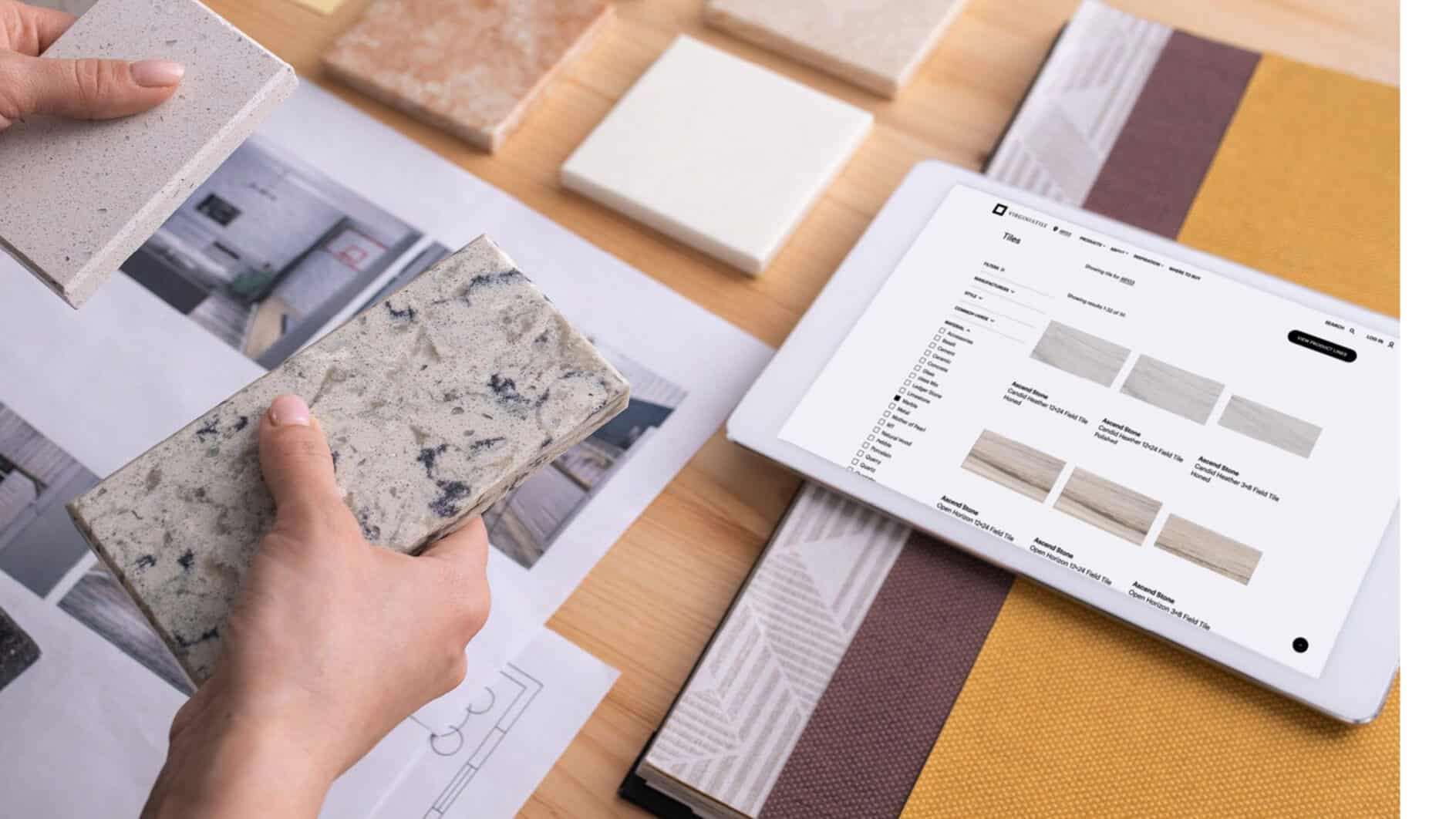 Hands hold stone samples over architectural plans on a wooden table. Nearby, a tablet displays a flooring website. Calm, professional atmosphere.