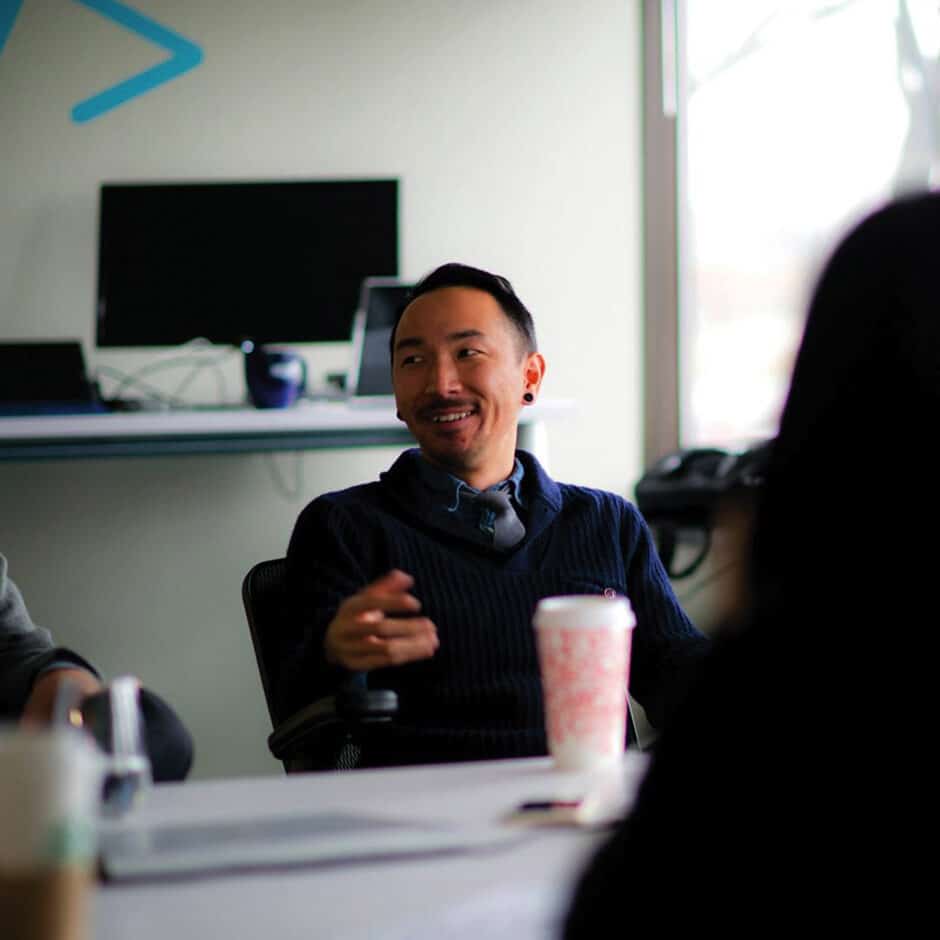 A man in a navy sweater, smiling and gesturing, sits at a table in a meeting room. A laptop is on a shelf behind him, creating a collaborative atmosphere.