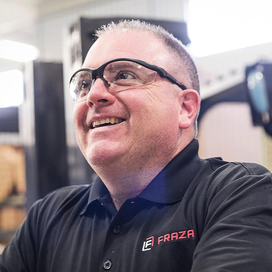 A smiling man in safety glasses and a black shirt with a "FRAZA" logo in a bright, industrial setting, conveying a positive, professional atmosphere.